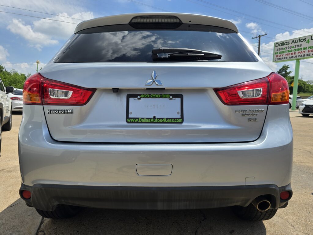 Rear view of a silver Mitsubishi Outlander Sport in a car dealership lot, with a Dallas Autos 4 Less sign in the background.