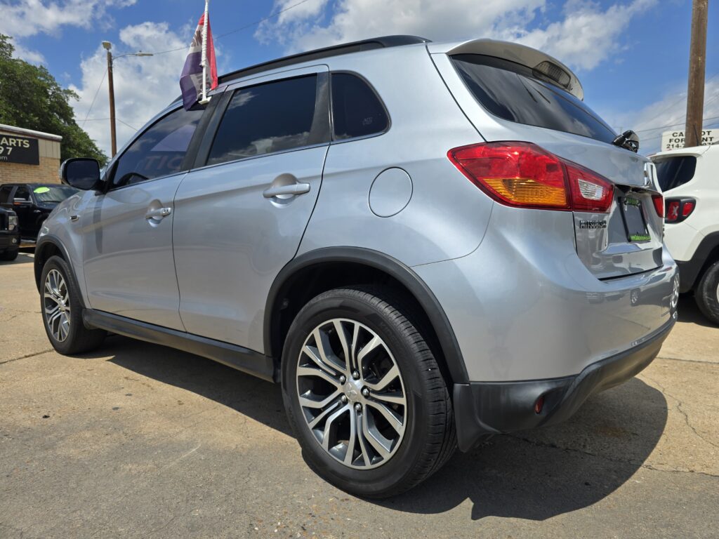 Silver Mitsubishi SUV seen from the rear-side, with a flag mounted on the roof, parked in a sunny car lot.