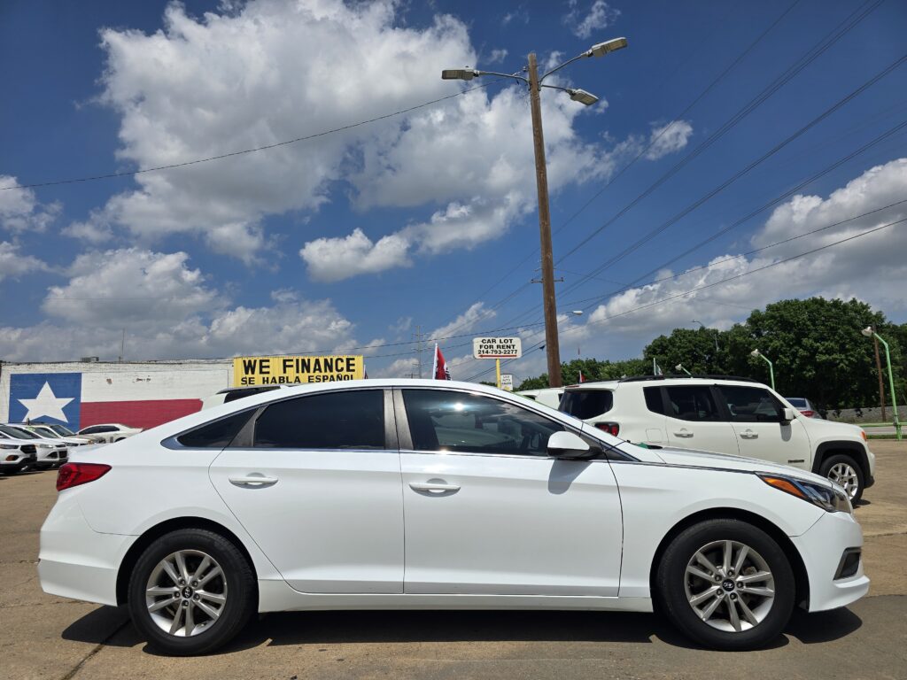 White SUV in a car dealership lot with a bold 'WE FINANCE' sign on a wall and a Texas flag mural, under a blue sky with clouds.