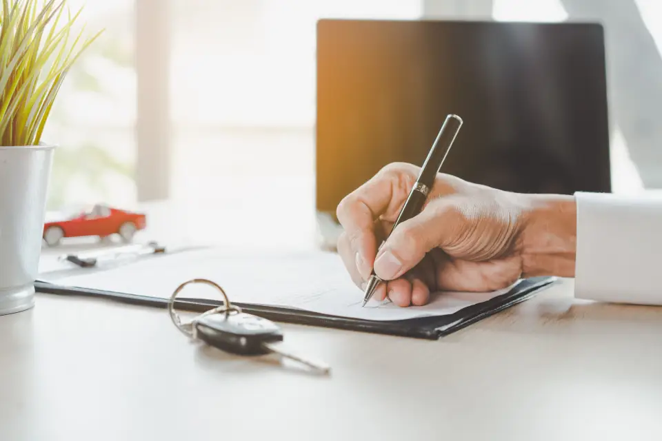 Close-up of a hand signing papers on a clipboard at a bright desk; car keys and a small red toy car nearby.