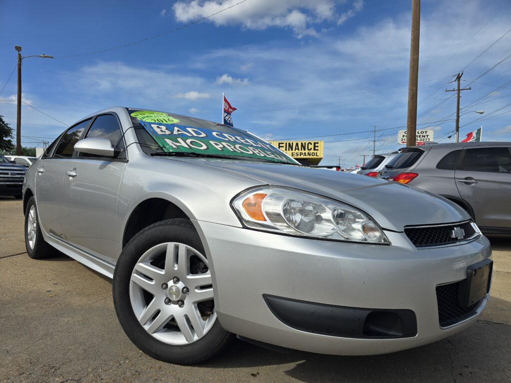 Silver sedan in a car lot with large green-on-black sign on the windshield reading 'BAD CREDIT NO PROBLEM' and other vehicles nearby under a blue sky with power lines and poles.