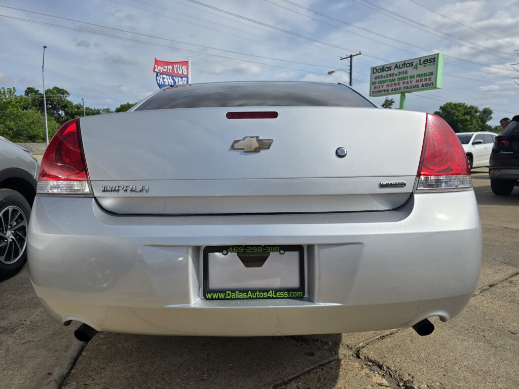 Rear view of a silver Chevrolet Impala on a car dealership lot, with a Dallas Autos 4 Less sign in the background and other cars around.