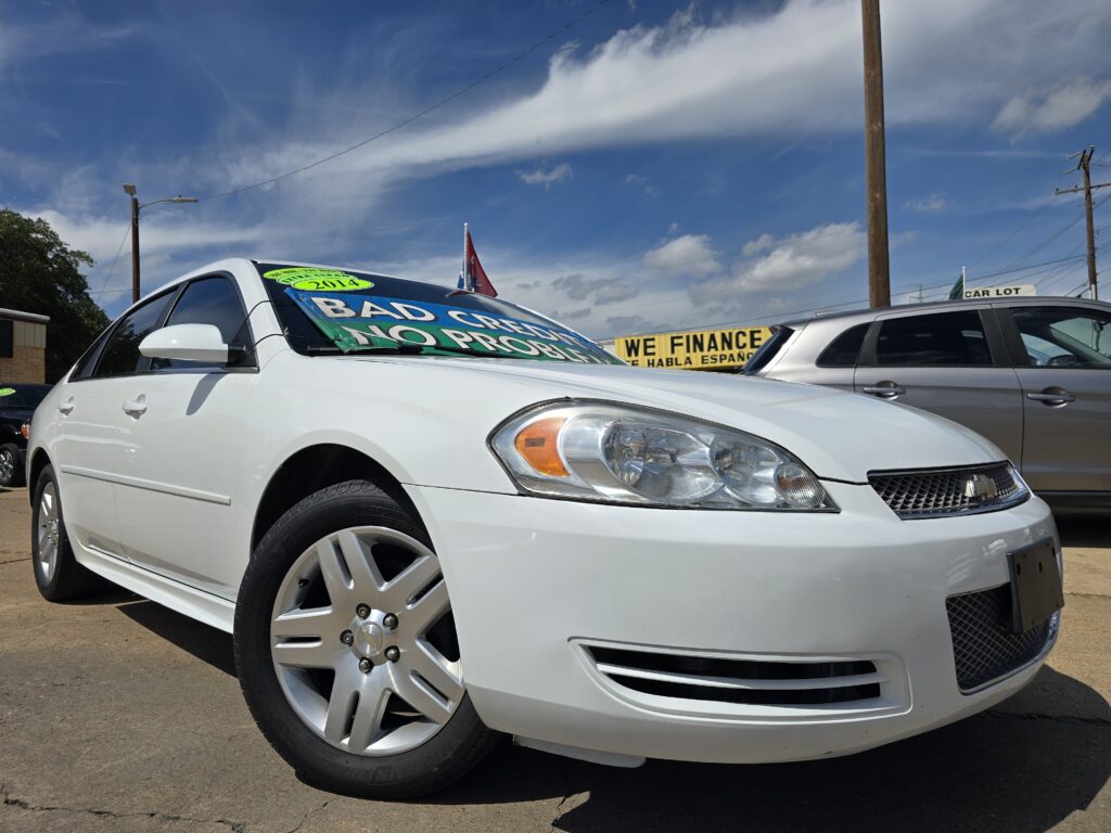 Close-up of a white car's front end in a dealership lot, with a bright windshield sign and clear blue sky above.