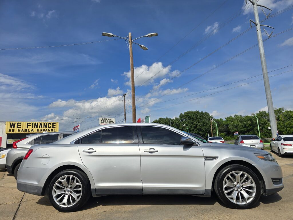 Silver sedan in foreground at a car dealership lot with a yellow WE FINANCE sign and blue sky above.