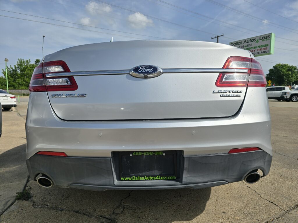 Rear view of a silver Ford Taurus with taillights, 'Taurus' badge, and 'Limited' badge in a parking lot; green Dallas Autos 4 Less sign in the background.