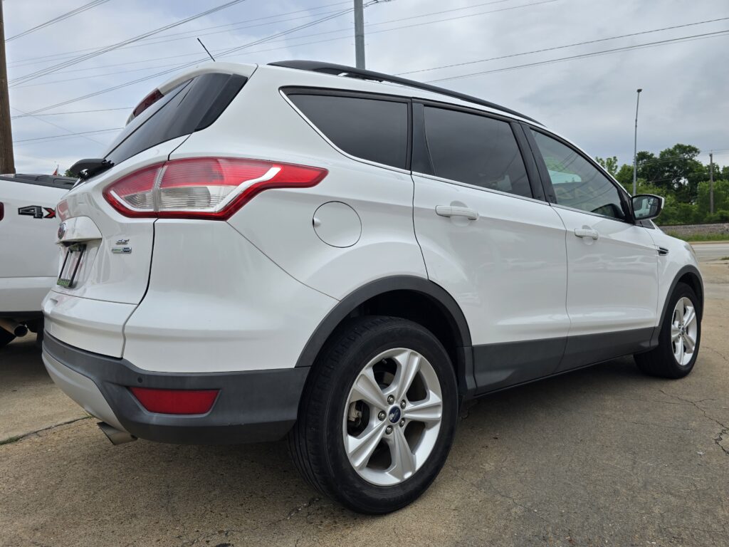 White SUV photographed from the rear-left side, showing the tail light, rear passenger door, and wheel on a paved lot with a pickup nearby.