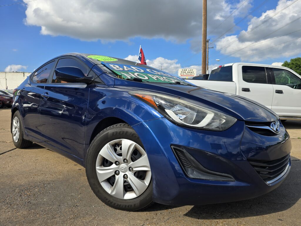 Blue Hyundai sedan in a dealership lot with a large windshield sign, white pickup behind, under a partly cloudy blue sky.