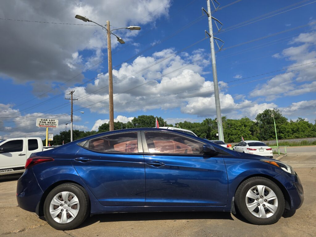 Blue SUV in a car lot with other vehicles, utility poles, and a blue sky with clouds behind it.