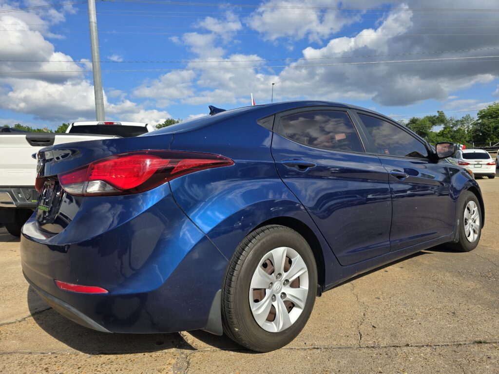 Blue sedan parked in a lot, seen from the rear-side with other vehicles and a cloudy blue sky above