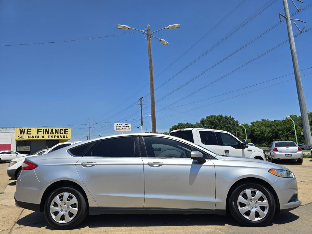 Silver sedan in the foreground of a car lot with a large yellow 'WE FINANCE' sign and other parked cars under a clear blue sky.