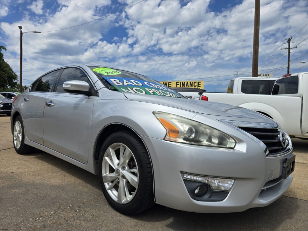 Side-angle view of a silver sedan in a car dealership lot with large colorful sale signs on the windshield, blue sky with clouds in the background, and other vehicles nearby.