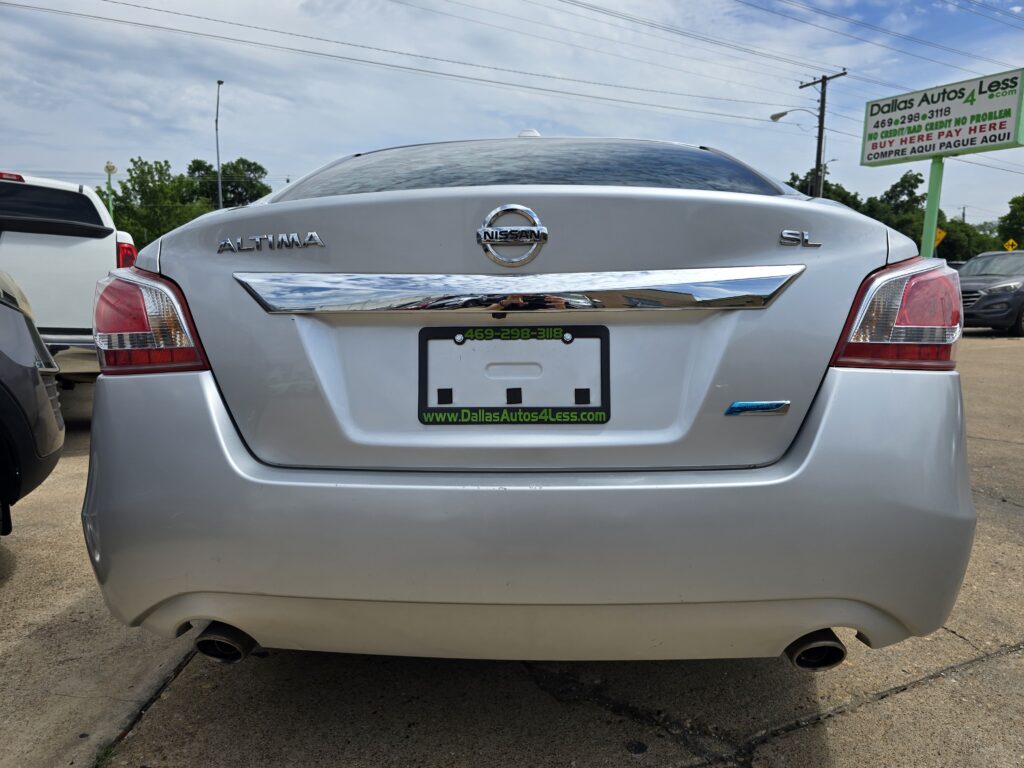 Rear view of a silver Nissan Altima SL in a car dealership lot, chrome bumper accent, and red taillights with a green-ad text plate frame reading Dallas Autos 4 Less in the background.