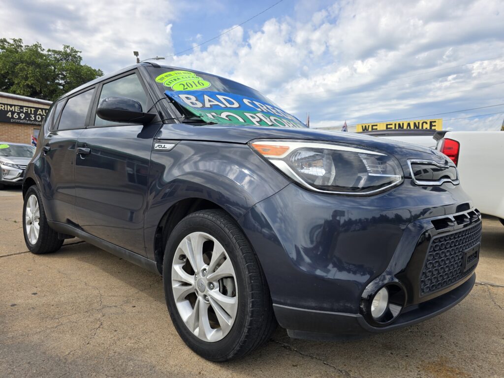 Dark gray minivan at a car dealership with a large sale banner across the windshield and a bright sky background.
