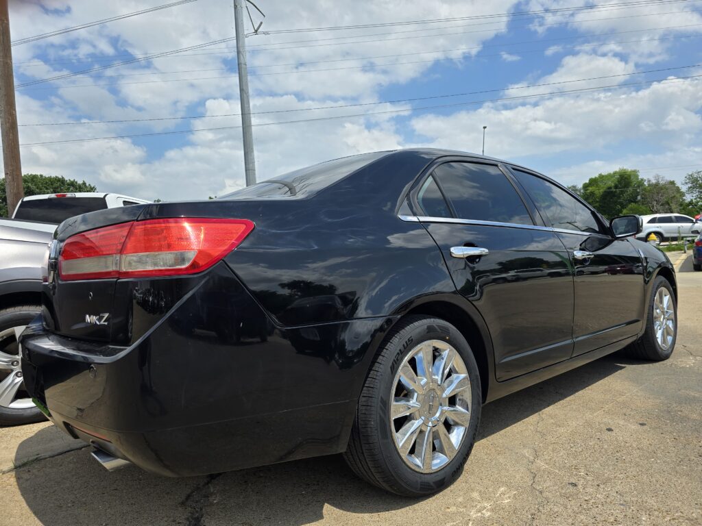 Close-up of a glossy black sedan's rear and side, chrome wheels, in a parking lot with other cars nearby.