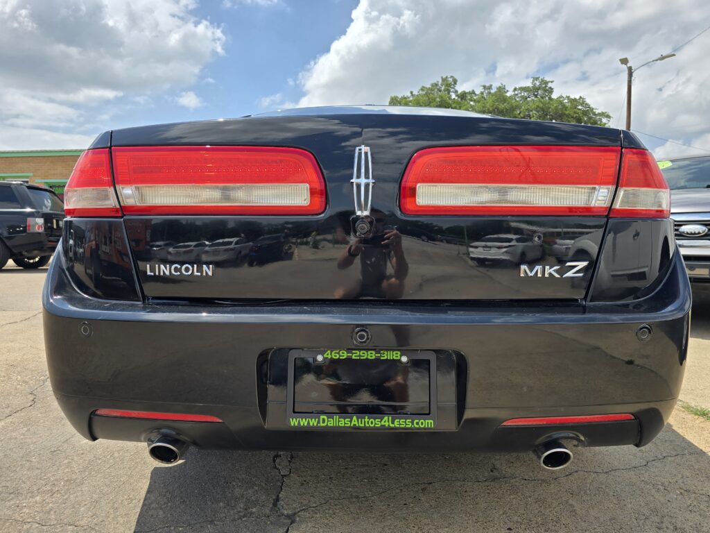 Rear view of a black Lincoln MKZ, showing red taillights, Lincoln emblem, and dealer plate frame in a parking lot.