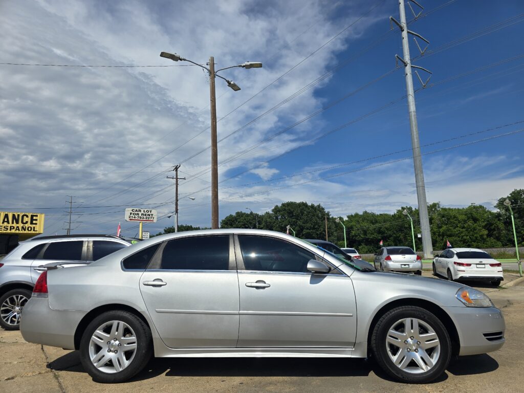 Row of silver cars parked in a car lot with utility poles and overhead power lines against a blue sky with clouds.