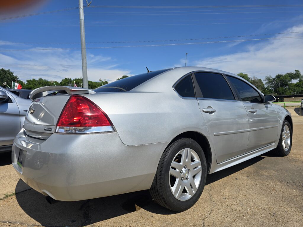Silver sedan parked in a car lot, showing its rear and right side with a spoiler and tinted windows under a blue sky.