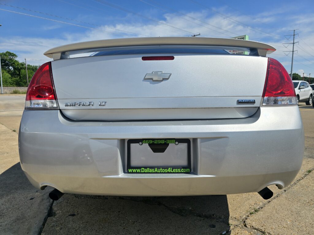 Rear view of a silver Chevrolet Impala LT with a beige spoiler in a parking lot, dealership plate frame showing a green DallasAutos4Less URL.