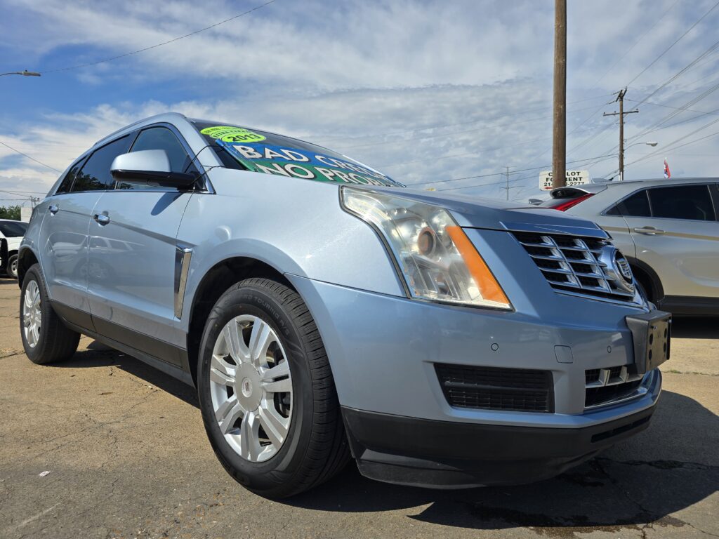 Silver Cadillac SUV on a car lot with large green sale signs visible in the windshield, parked beside other vehicles under a blue sky.