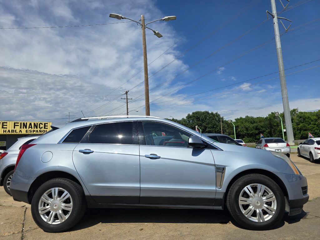 Silver Cadillac SUV parked in a car dealership lot with a bright yellow sign advertising financing in the background and power lines overhead against a blue sky.