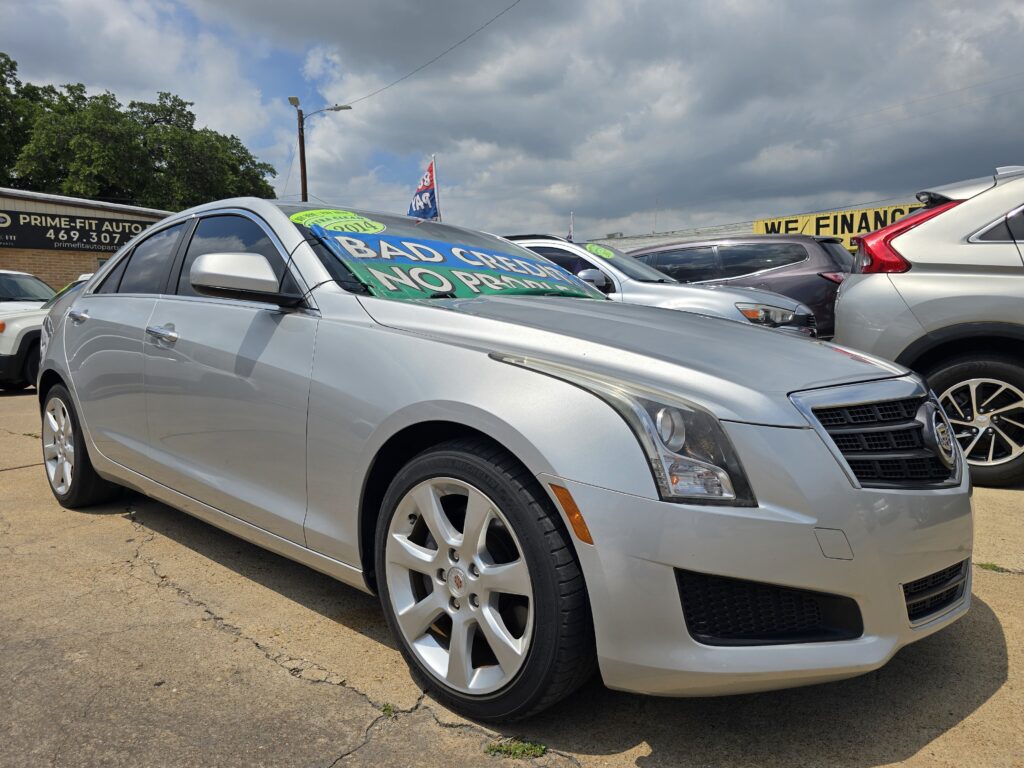 Silver Cadillac in a dealership lot with large sale signs on the windshield and a 'WE FINANCE' banner in the background, under a cloudy sky.