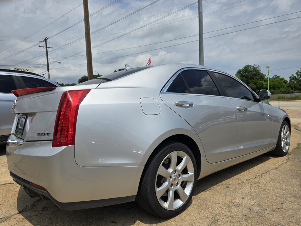 Silver Cadillac sedan viewed from the rear side in a parking lot with power lines and a cloudy sky above.