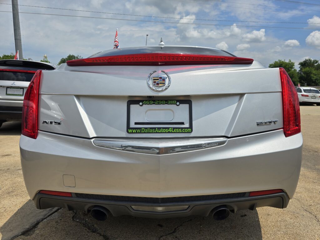 Back of a silver Cadillac SUV with red taillights and a green dealership plate frame reading www.DallasAutos4Less.com in a car lot.