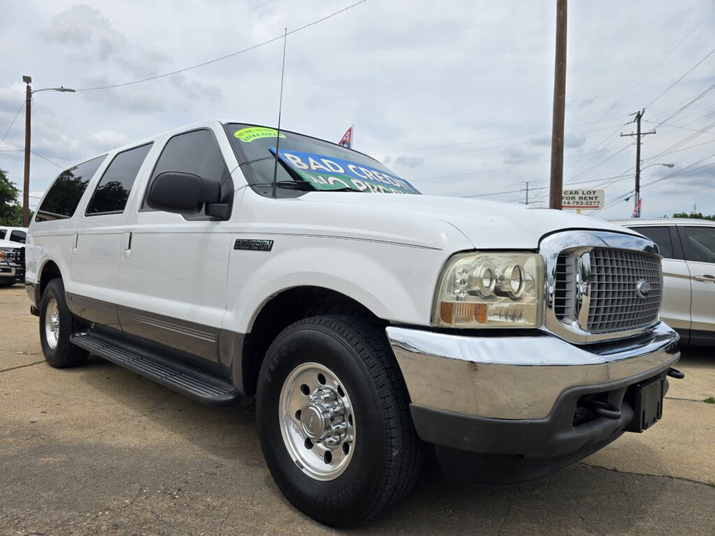 White Ford Excursion van with a bright blue 'BAD CREDIT' sign on the windshield at a car dealership lot.