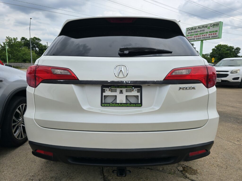 Rear view of a white Acura SUV (RDX) showing taillights, rear windshield wiper, and a hitch, with a dealership plate frame reading Dallas Autos 4 Less.