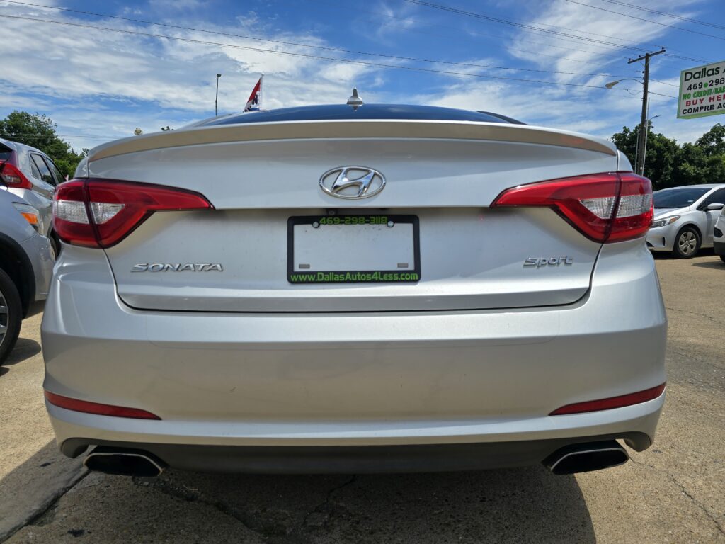 Rear view of a silver Hyundai Sonata Sport in a car dealership lot, with a dealer plate frame reading www.DallasAutos4Less.com and a 469-298-3118 contact number visible on the plate area against a blue sky.