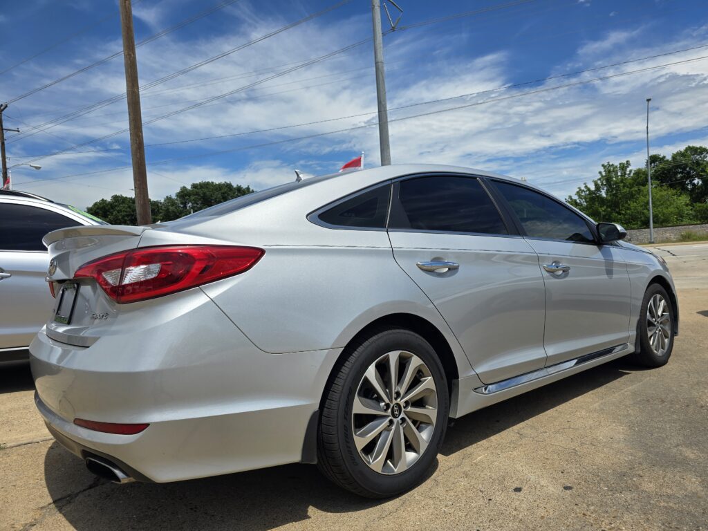 Silver Hyundai Sonata parked on a curb, showing rear and side profile with another car to its left and a bright blue sky above.