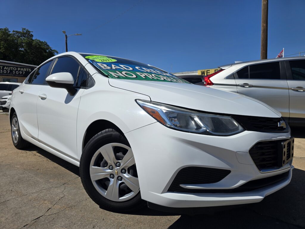 White sedan in a car dealership lot with bright green sale signs on the windshield against a clear blue sky.