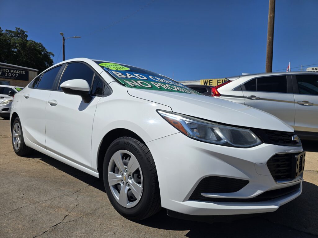 White sedan parked at a used-car lot with a large green sale sign on the windshield saying 2019 or similar year options visible in bold letters.