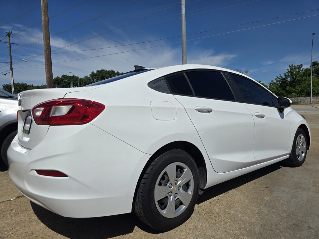 White sedan parked in a lot, rear quarter panel and taillight visible against a clear blue sky.