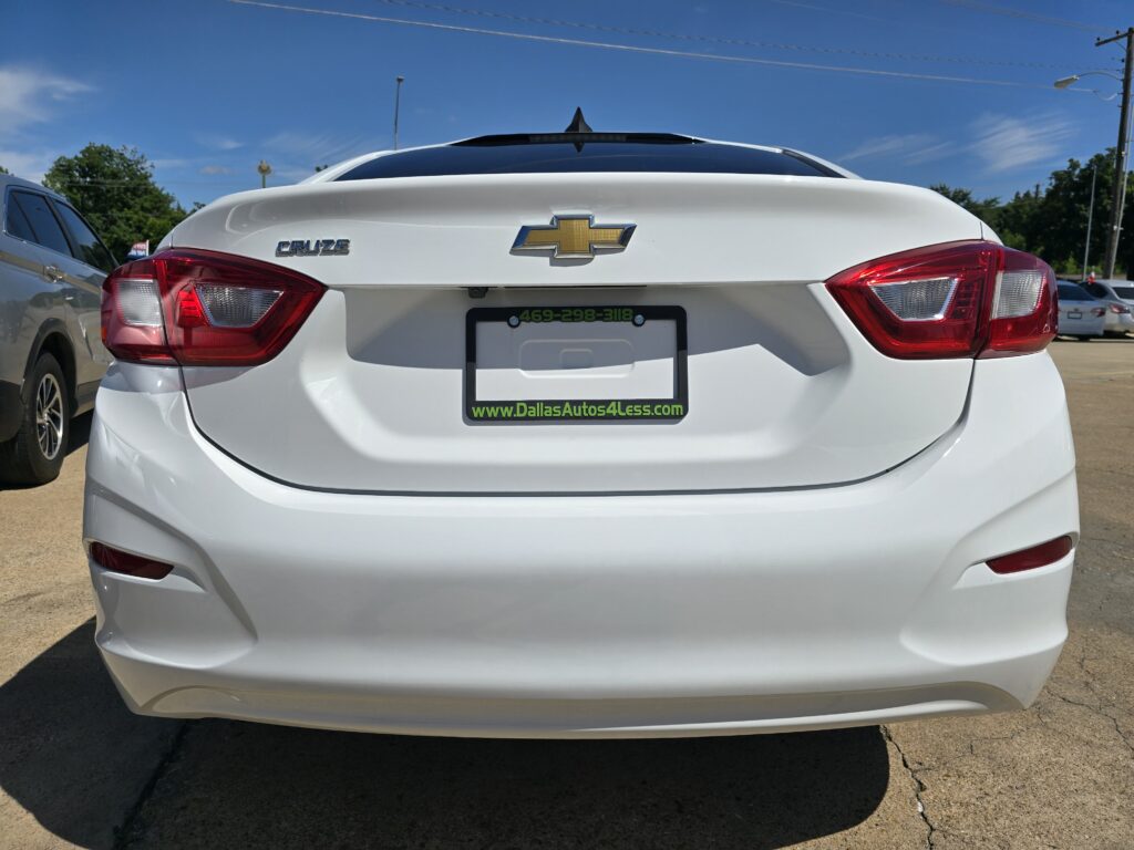 Rear of a white Chevrolet Cruze showing the Chevrolet bowtie logo, Cruze badge, red tail lights, and a green Dallas Autos 4 Less license frame in a parking lot.