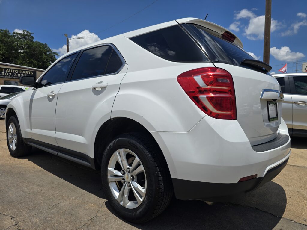 White SUV parked at a car dealership lot, showing the rear three-quarter view with red taillights and tinted rear windows under a blue sky.