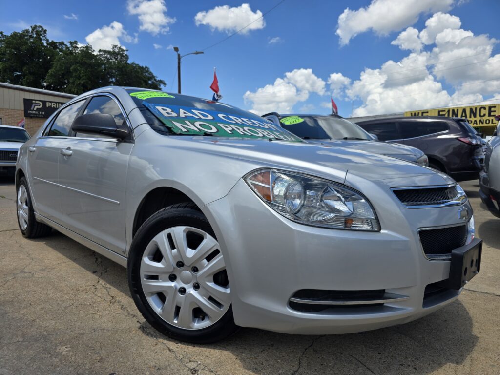 Close-up of a silver Chevrolet sedan in a car dealership lot, windshields covered with green sale banners about bad credit and financing.