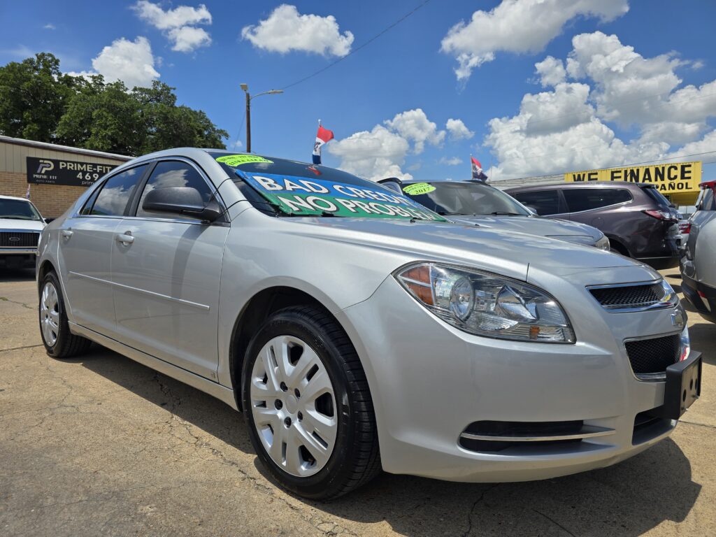 Front-left view of a silver sedan at a car dealership, with bright green promotional signs on the windshield and a 'We Finance' sign in the background.