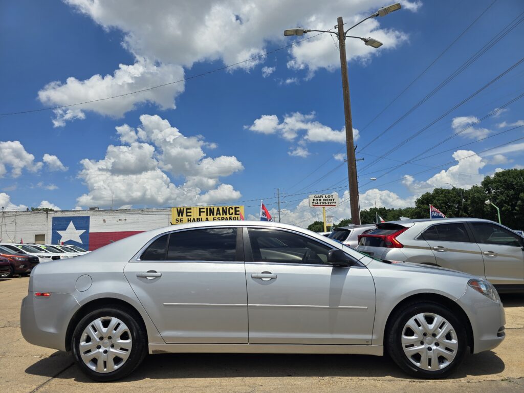 Parking lot with row of cars, yellow 'WE FINANCE' sign and Texas flag mural on wall left under blue sky with clouds.