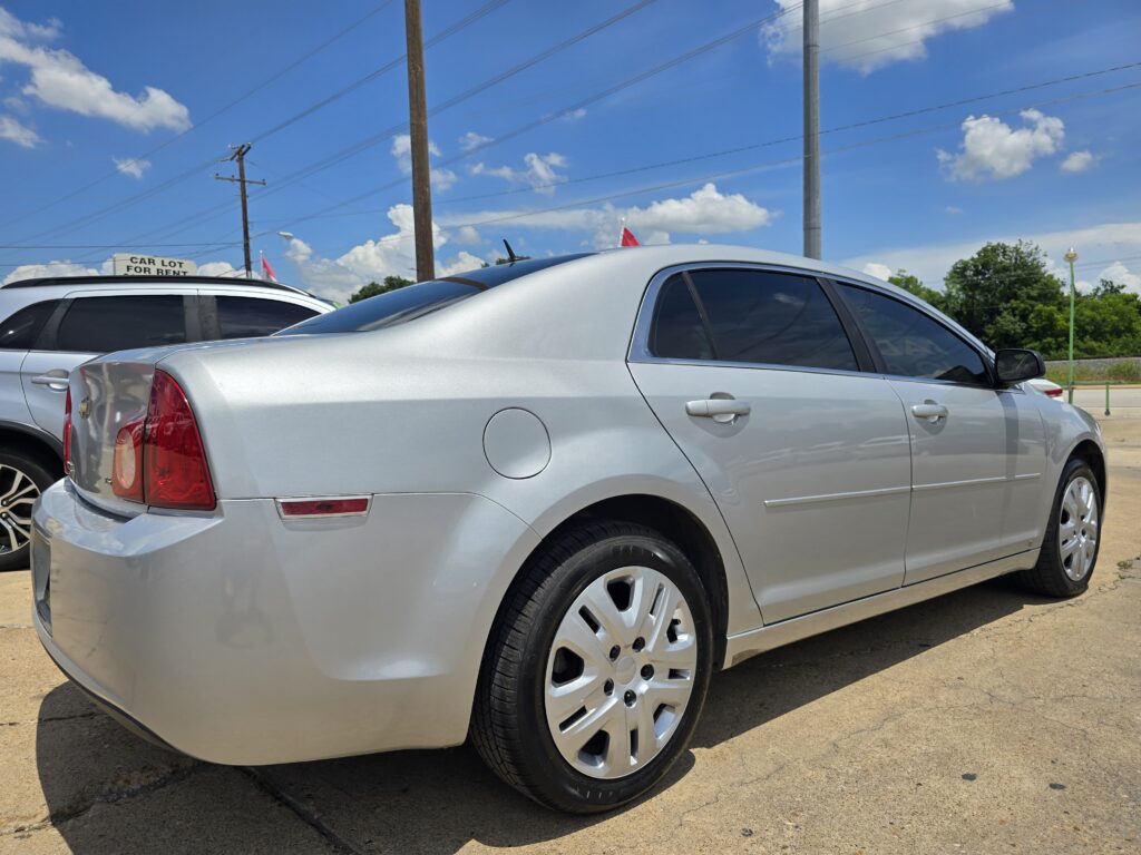 Close-up view of a silver sedan in a car lot, with another car beside it and a blue sky above with power lines in the background.