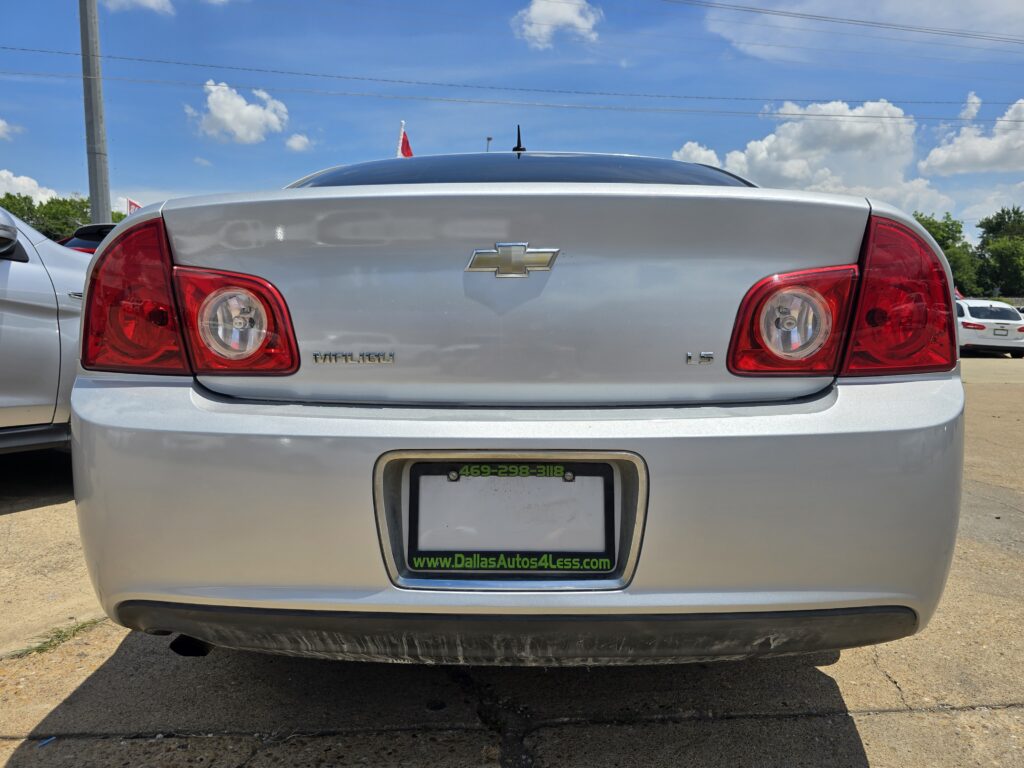 Rear view of a silver Chevrolet Malibu LS in a parking lot, showing the Chevy emblem, red taillights, and a Dallas Autos 4 Less license plate frame under a bright blue sky.