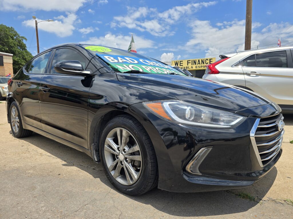 Black sedan in the foreground with owner-placed sale signs on the windshield; other cars and a 'We Finance' sign visible in the background under a blue sky.
