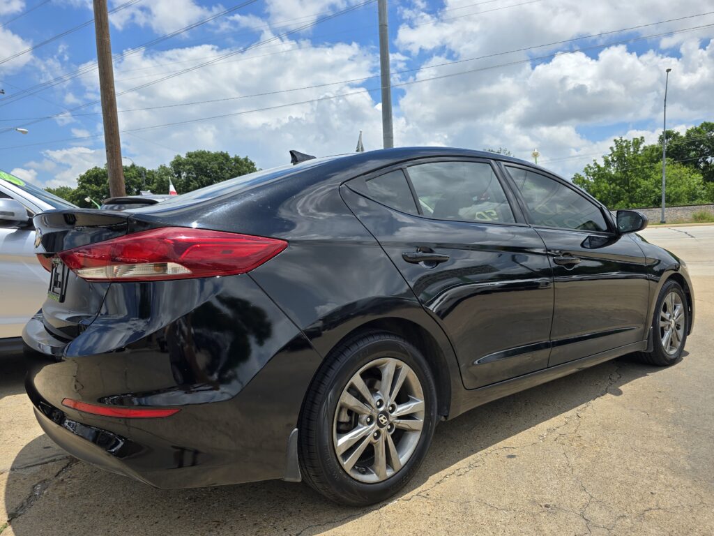 Rear-left view of a shiny black sedan with a subtle spoiler in a parking lot under a blue sky with clouds