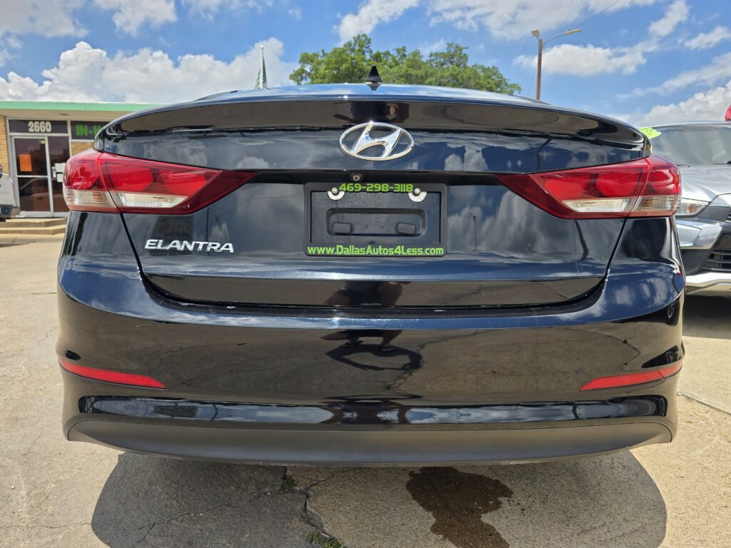 Rear view of a black Hyundai Elantra at a car lot, with a green phone number and Dallas Autos 4 Less frame on the license area.