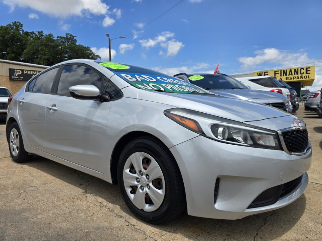 Silver sedan in a car dealership lot with a bright windshield banner and a 'WE FINANCE' sign visible in the background on a sunny day, blue sky.