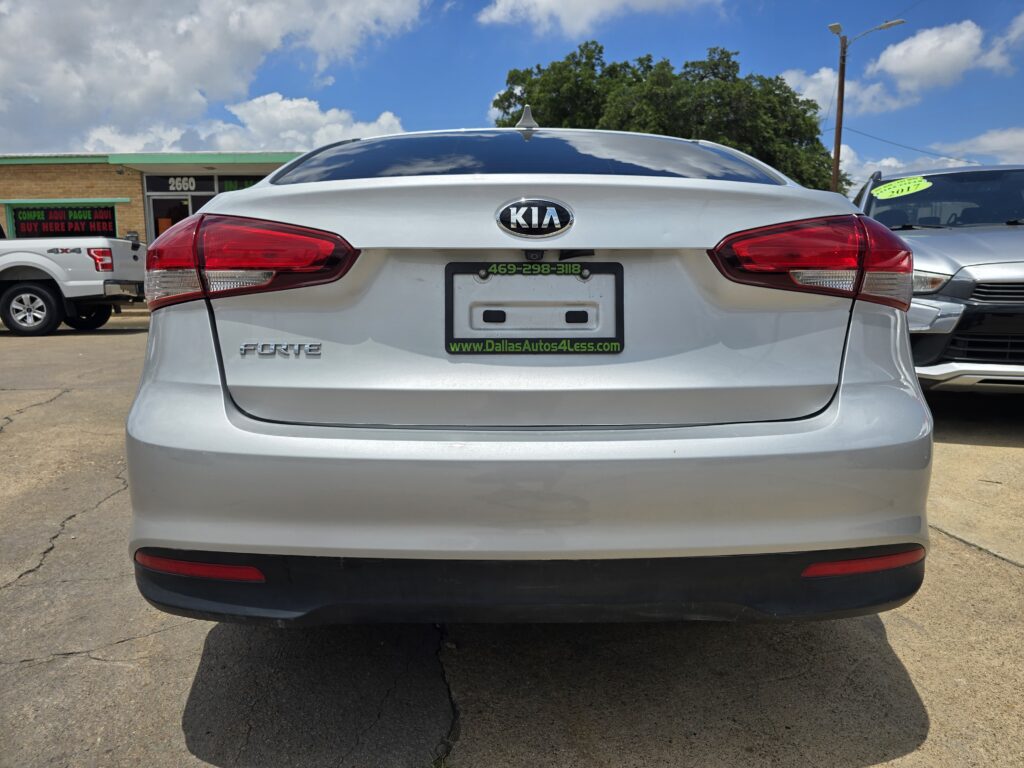Rear view of a silver Kia Forte in a car lot, dealership signs in the background.