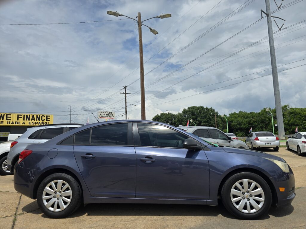 Line of cars parked at a dealership lot with a bright yellow 'WE FINANCE' sign in the foreground and overhead power lines and poles under a cloudy sky.