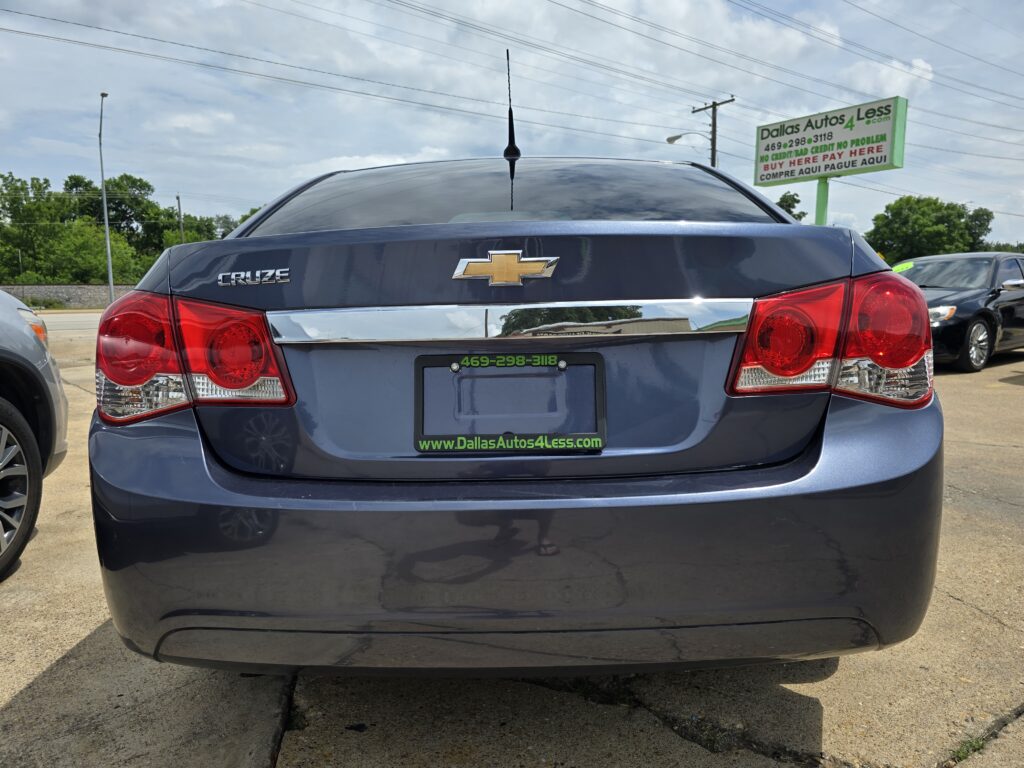 Rear view of a dark blue Chevrolet Cruze with Chevrolet bowtie emblem, shown in a car lot with a green Dallas Autos 4 Less sign in the background.