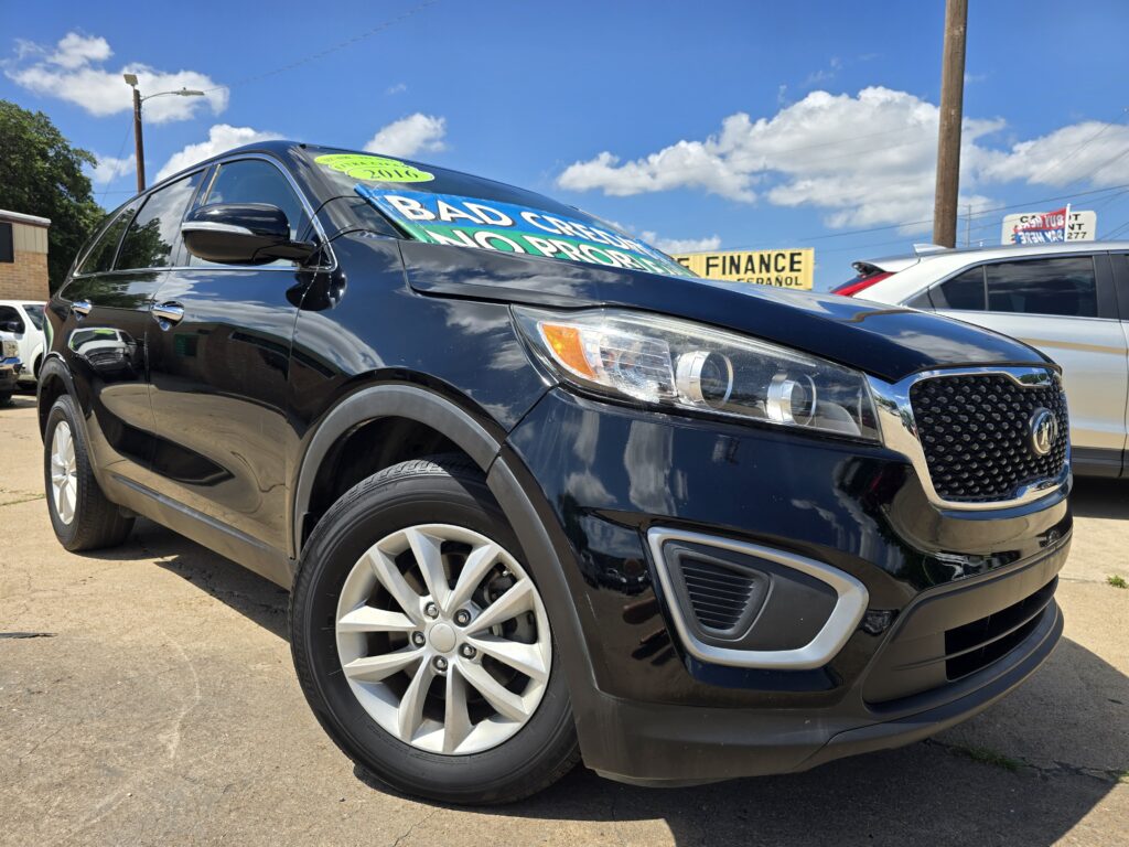 Black SUV on a dealership lot with large sale signs on the windshield under a blue sky with clouds