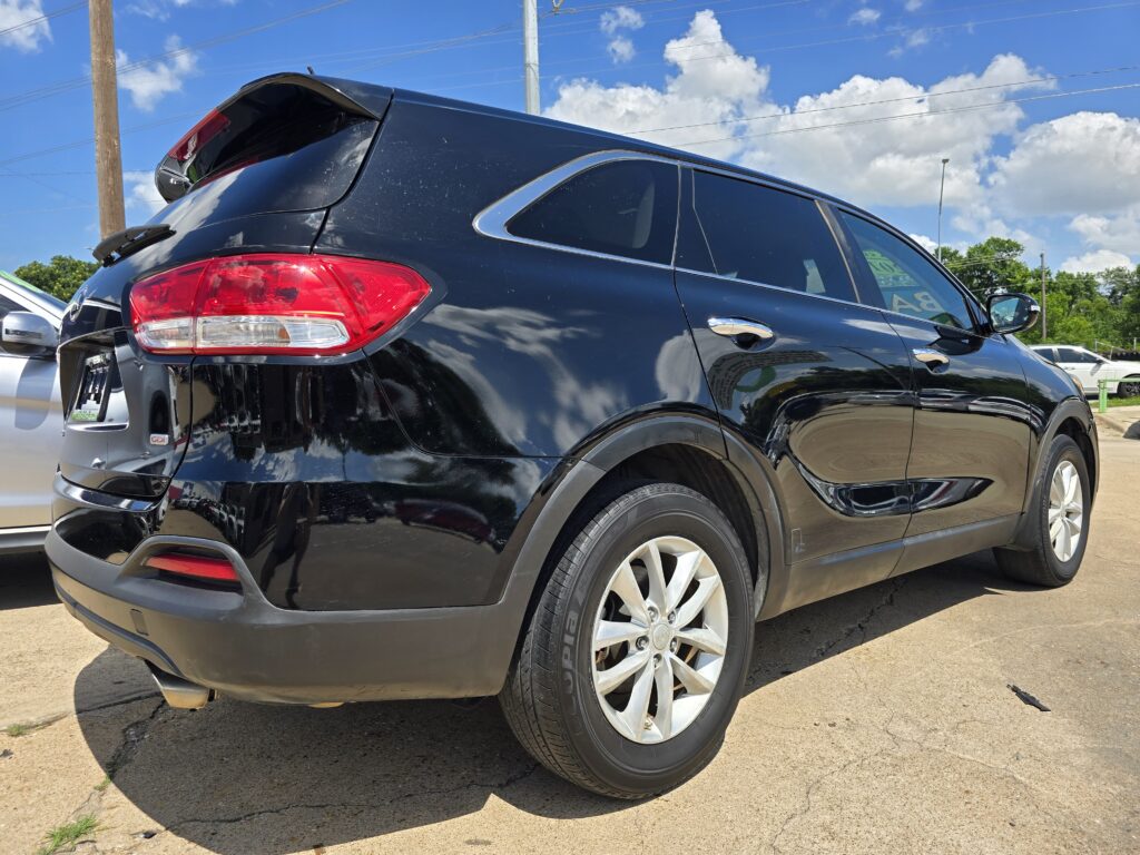 Rear three-quarter view of a black SUV parked on a paved lot, showing pronounced taillight, alloy wheel, and small open tailgate area against a blue sky.